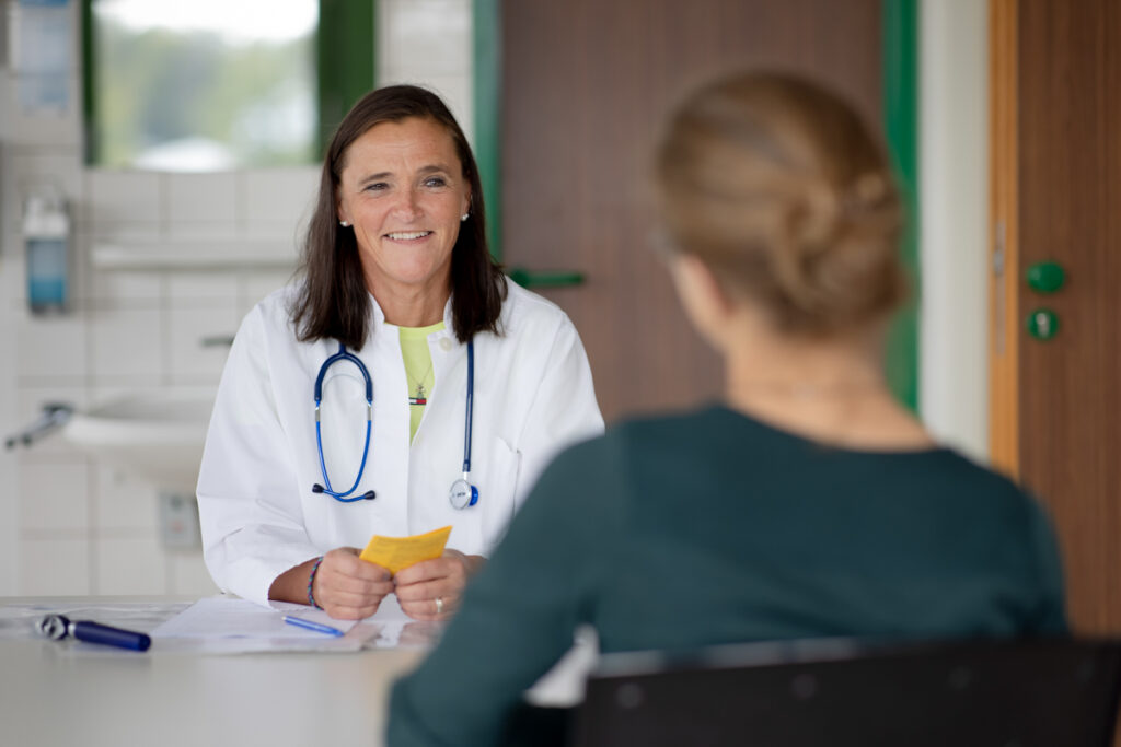 Eine Ärztin in weißem Kittel und Stetoskop um den Halz lächelt.