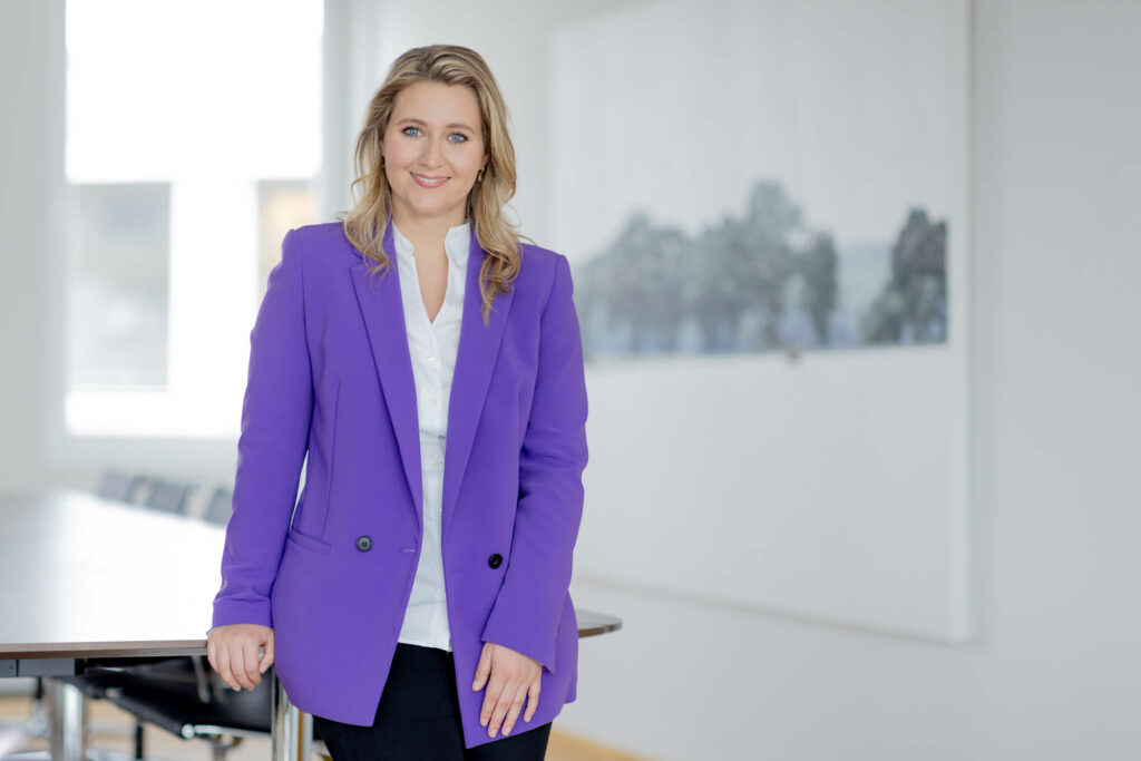 Frau mit blonden langen Haaren, weißer Bluse, lila Blazer steht an einem Tisch angelehnt und lächelt.