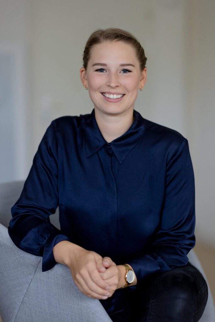 Frau mit blonden Haaren und blauer Bluse sitzt auf einem Sessel. Sie lacht.