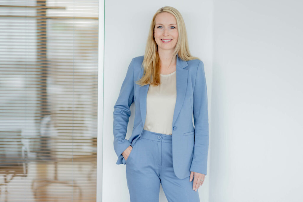 Frau mit langen blonden Haaren, weißer Bluse und hellblauem Hosenanzug. Ihre rechte Hand ist in der Hosentasche und sie steht vor einer weißen Wand.