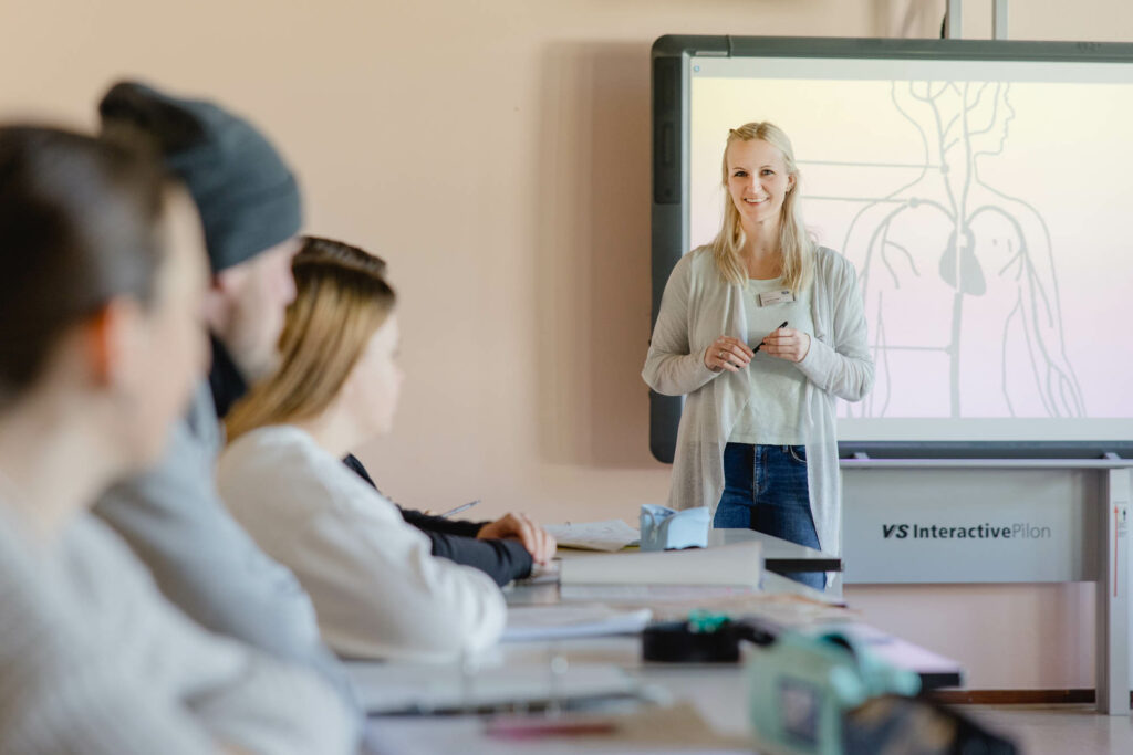 Eine Frau mit blonden Haaren steht for einem Smartboard und hält einen Stift in der Hand. Rechts sitzen Schülerinnne und Schüler an Tischen.