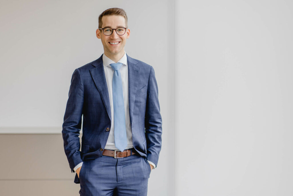 Mann mit kurzen dunklen Haaren, Brille, in blauem Hosenanzug und hellblauer Krawatte. Er hat die Hände in den Hosentaschen und lächelt.