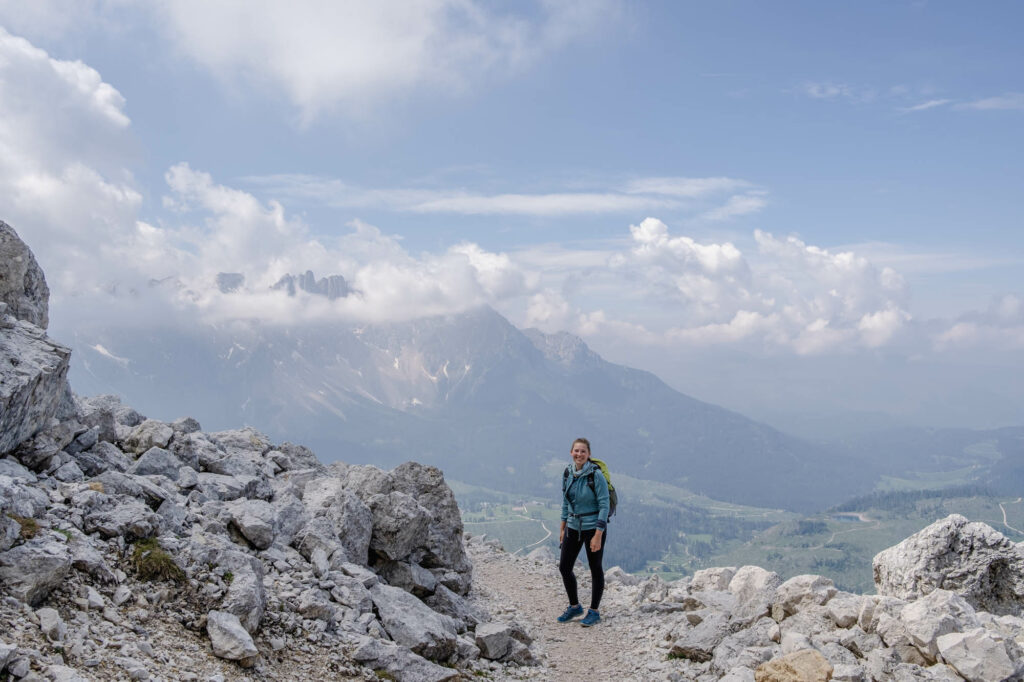 Gebirgslandschaft im Hintergund, Frau mit blonden Haaren steht in Wanderng auf einem Schotterweg.