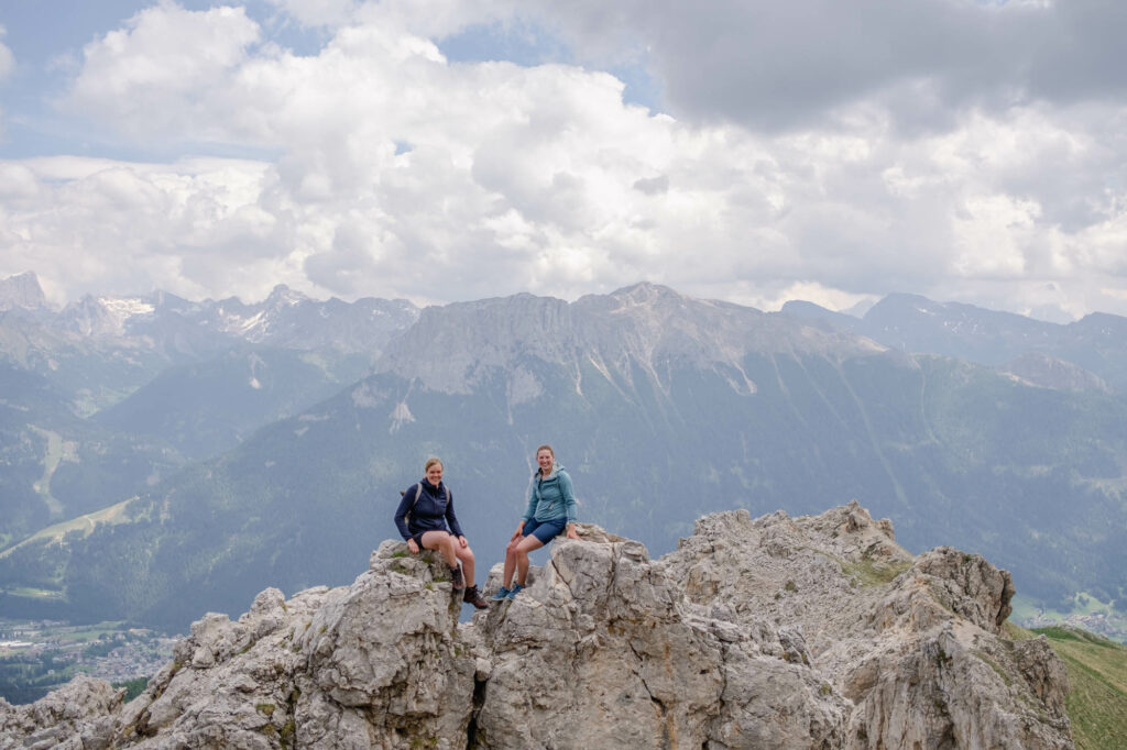 Zwei Frauen sitzen auf Felsen. Im Hintergund sieht man Berge und Wolken.
