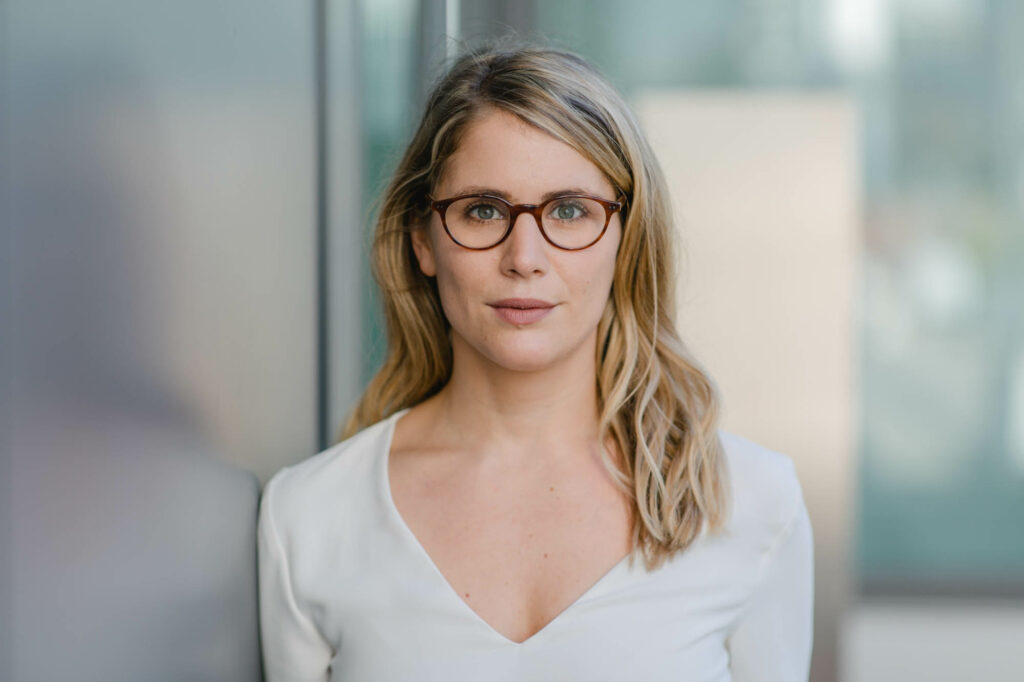 Frau mit langen blonden Haaren. Sie trägt beige Bluse und dunkle Brille.