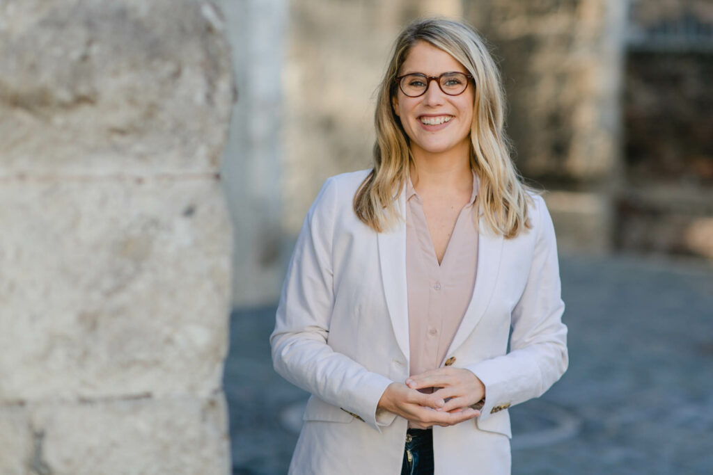 Frau mit langen blonden Haaren. Sie trägt weißen Blazer, beige Bluse und dunkle Brille. Sie lacht.