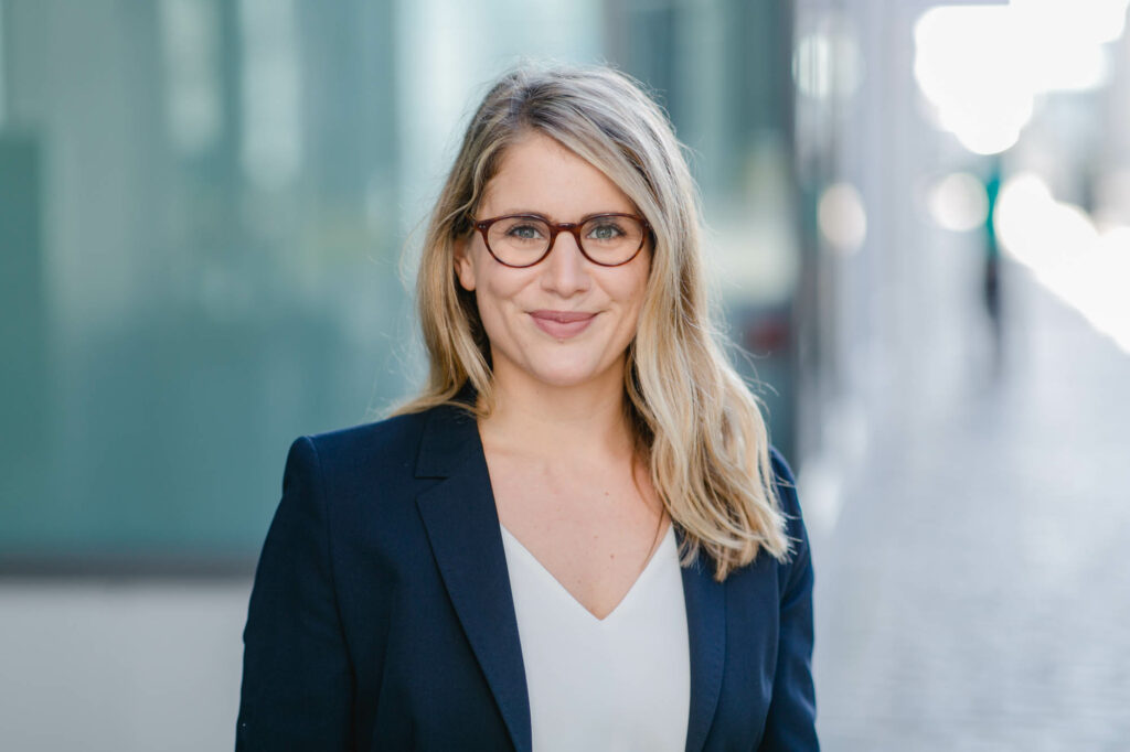 Frau mit langen blonden Haaren. Sie trägt blauen Blazer, beige Bluse und dunkle Brille. Sie lächelt..