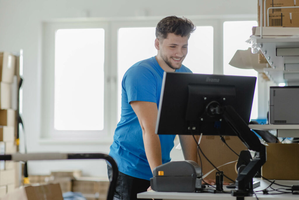 Mann mit kurzen dunklen Haaren, Bert und blauem T-Shirt steht in einem Lagerraum vor einem Computer. Er lacht.