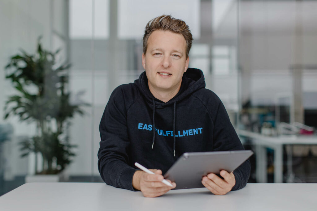 Mann mit kurzen blonden Haaren, schwarzen Pullover sitzt an einem Tisch. Er hät ein Tablet in der Hand.