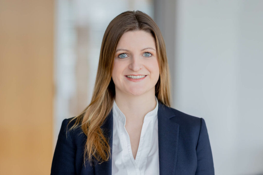 Frau mit braunen langen Haaren lacht. Sie trägt eine weiße Bluse und einen blauen Blazer.