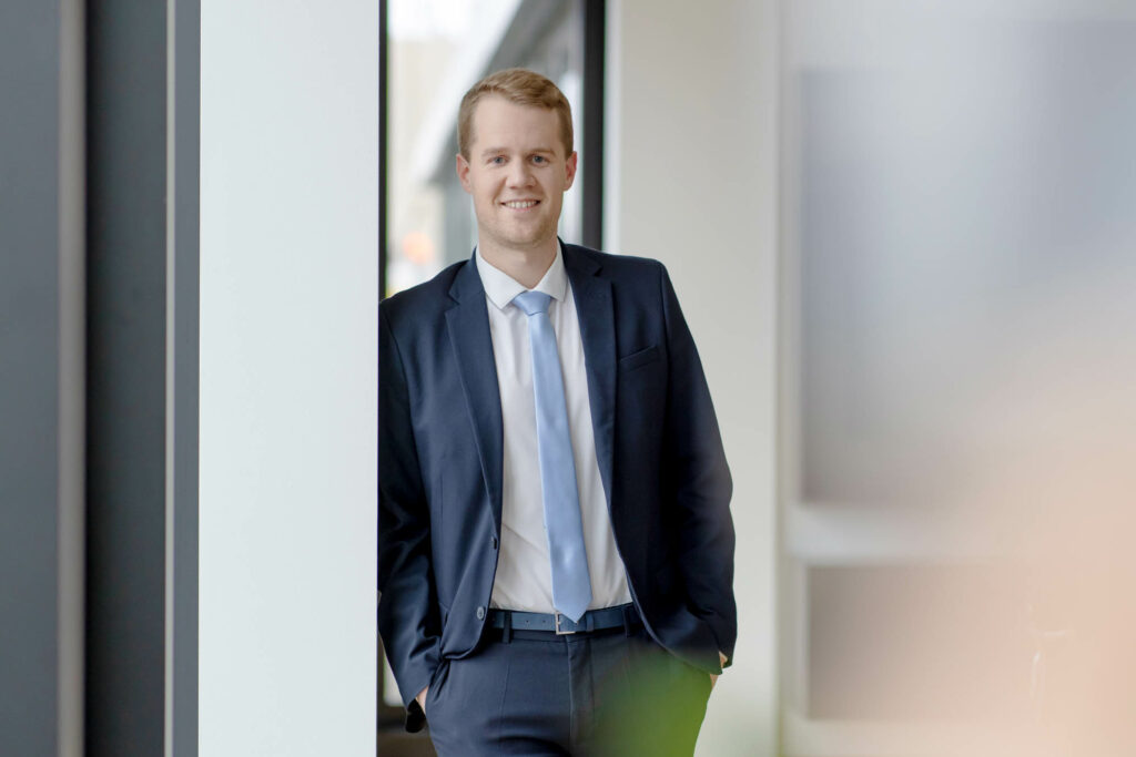 Mann mit blonden kurzen Haaren leht links an euner Wand. Er trägt einen blauen Anzug und eine hellblaue Krawatte und lächelt.