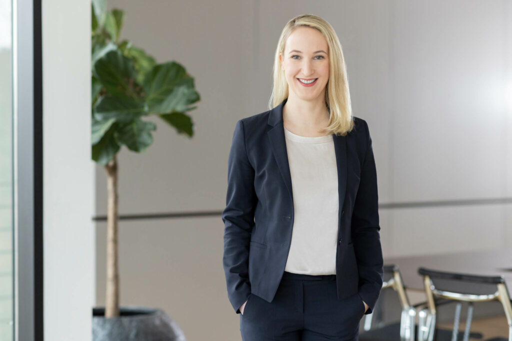 Frau mit blonden, langen Haaren trägt einen dunklen Hosenanzug und eine weiße Bluse. Sie lacht. Links im Hintegrund steht eine Grünplanze.