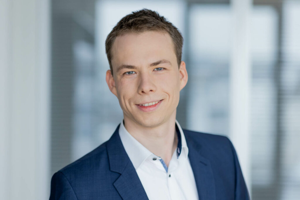 Portrait eines Mannes mit kurzen dunklen Haaren. Er lacht und trägt ein weißes Hemd und ein blaues Jackett.