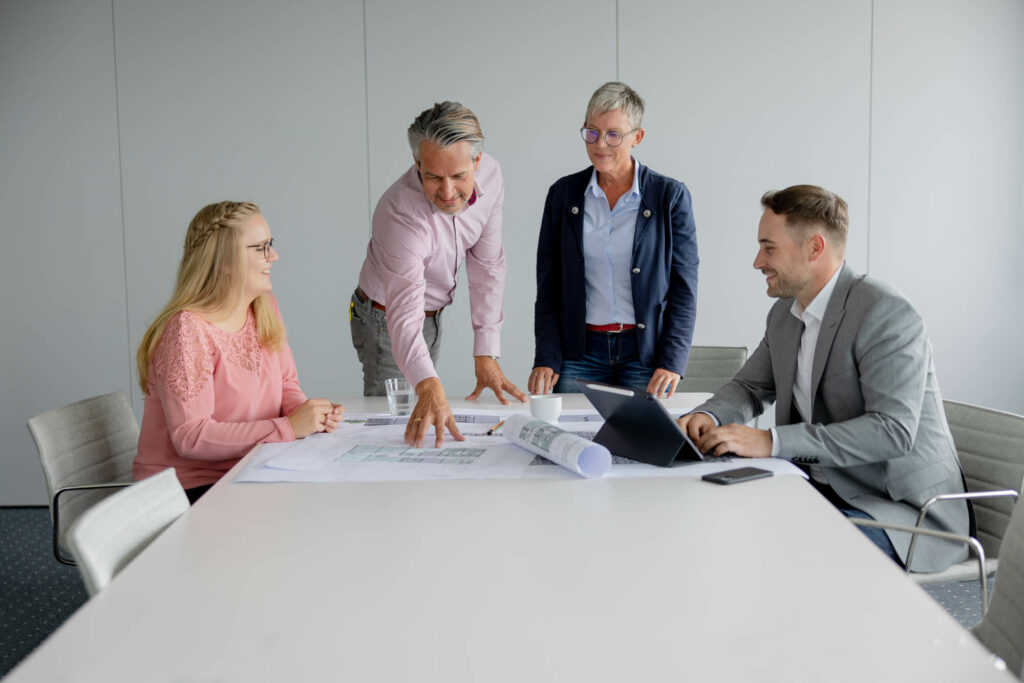Eine blonde Frau sitzt links an einem Schreibtisch. Ihr gegenüber sitzt ein Mann mit einem Laptop. Zwischen ihnen stehen zwei Männer. Sie unterhalten sich. Pläne liegen auf dem Tisch.