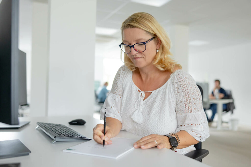 Eine blonde Frau sitzt an einem Schreibtisch und schreibt etwas auf einen Block. Sie trägt eine weiße Bluse und eine dunkle Brille.