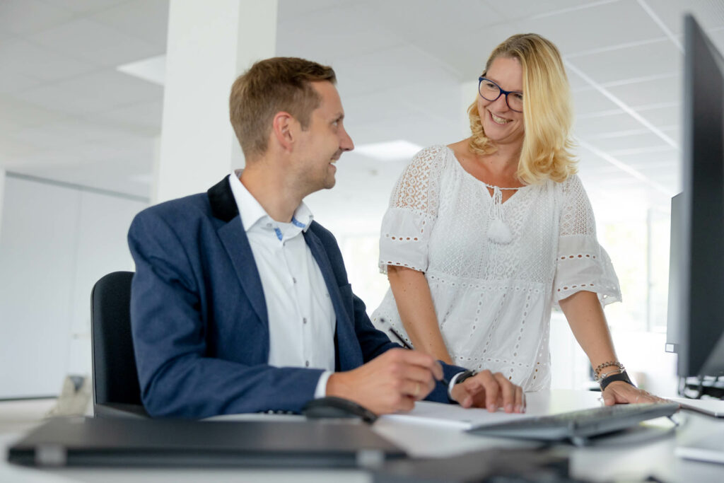 Ein Mann sitzt an einem Schreibtisch. Rechts neben ihm steht eine blonde Frau. Sie reden mit einander und lächeln.
