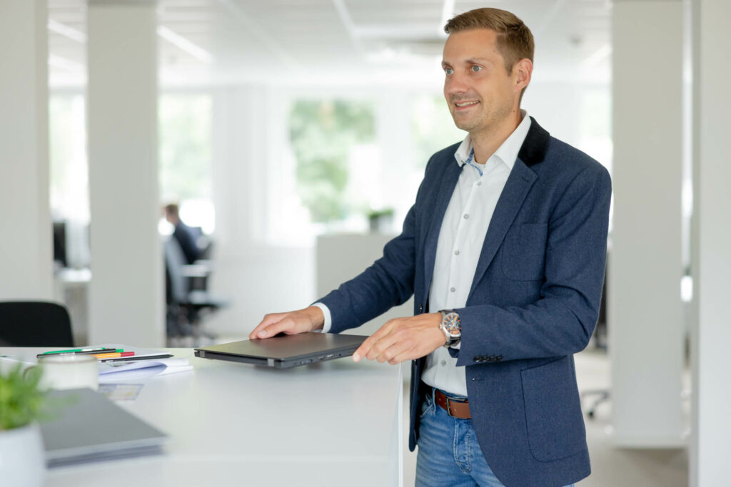 Mann mit blonden kurzen Haaren steht an einem Stehtisch und hält einen Laptop in der Hand. Er trägt ein blaues Jackett und lächelt.