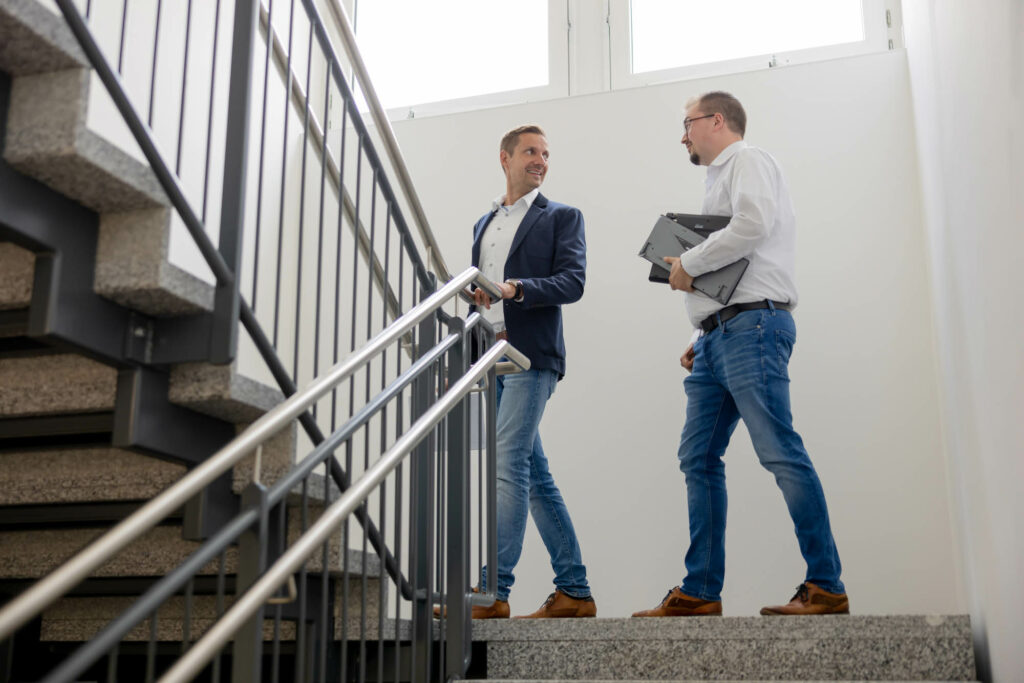 Zwei Männer stehen in einem Treppenhaus und unterhlaten sich. Ein Mann trägt Akten unter dem Arm.