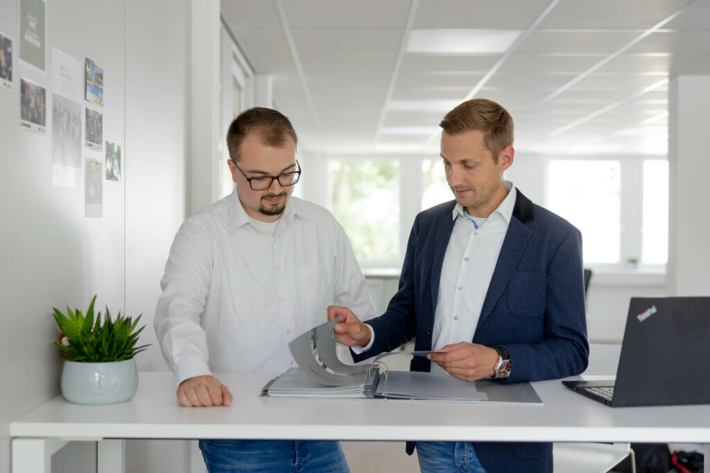 Zwei Männer stehen an einem Stehtisch. Sie blättern inm einem Magazin. Rechts steht ein Laptop.