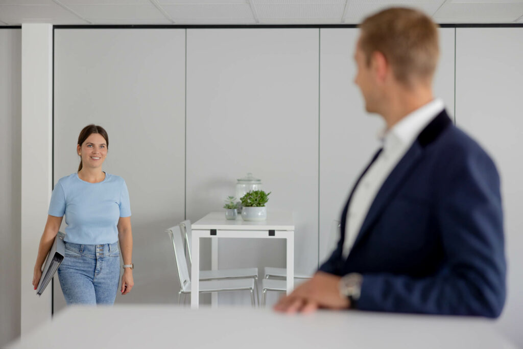 Frau mit dunklen Haaren, nach hinten gebunden trägt ein hellblaues T-Shirt, Jeand, trägt einen Aktenordner unter dem Arem und läuft auf einen Mann rechts zu, der an einem Regal lehnt. Er hat blonde Haaren uns trägt einen blauen Anzug.