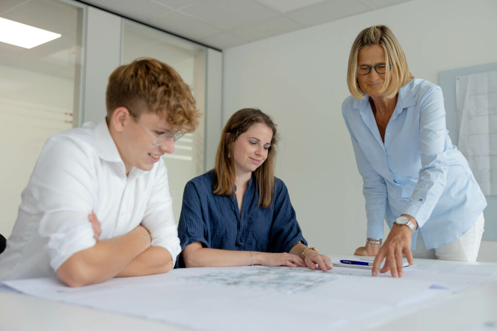 Mann mit weißem Hemd und blonden Locken sitzt links an einem Tisch. Rechts neben ihm eine Frau mit dunklen langen Haaren. Sie schauen auf einen Plan. Rechts steht eine Frau mit blonden, schulterlangen Haaren und zeigt etwas auf dem Plan.