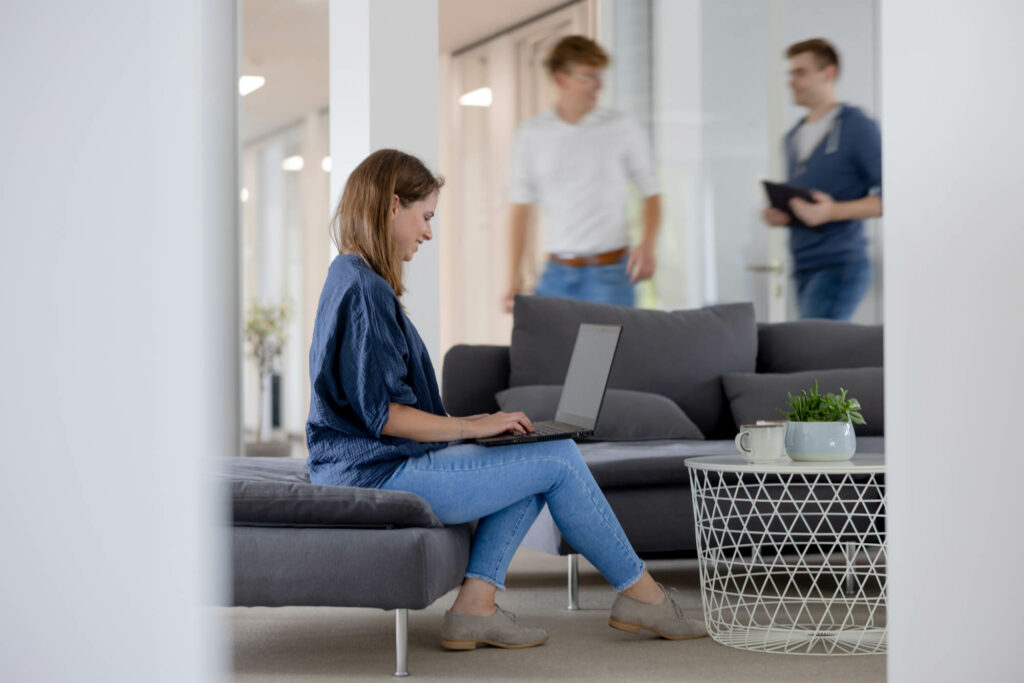 Frau mit dunklen Haaren sitzt auf einer Couch. SIe trägt eine blaue Bluse und Jeans, ein Laptop auf ihrem Schoß. Im Hintegrund laufen zwei Personen.