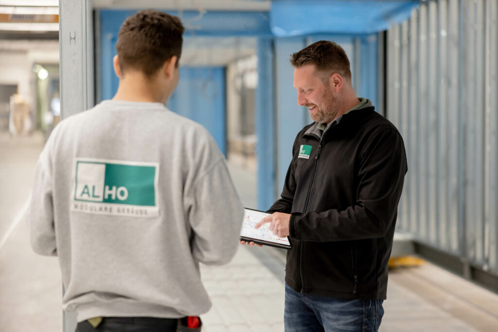 Zwei Männer stehen in einer Industriehalle. Links im Bild ein Mann mit grauem Pulli und grünem ALHO-Logo. Rechts daneben ein Mann in schwarzer Jacke. Er tippt auf einem Tablet.