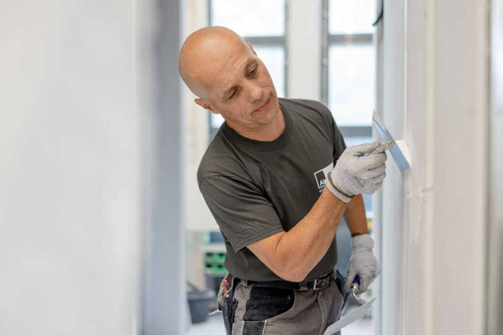 Mann in grauem T-Shirt und Glatze spachtelt eine Wand.