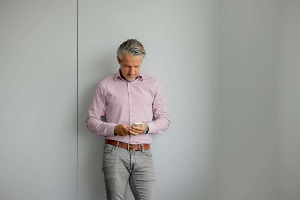 Mann mit grauen kurzen Haaren, rosa Hemd, grauer Hose steht vor einer hellen Wand. Er schaut auf ein Handy in seiner Hand.