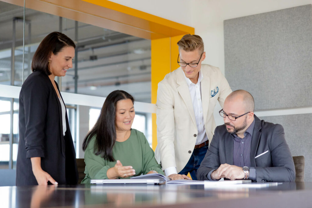 Vier Personen sind in einem Büro. Eine Frau und ein Mann sitzen an einem Tisch. Danaben stehen zwei Personen. Sie schauen auf Unterlagen.