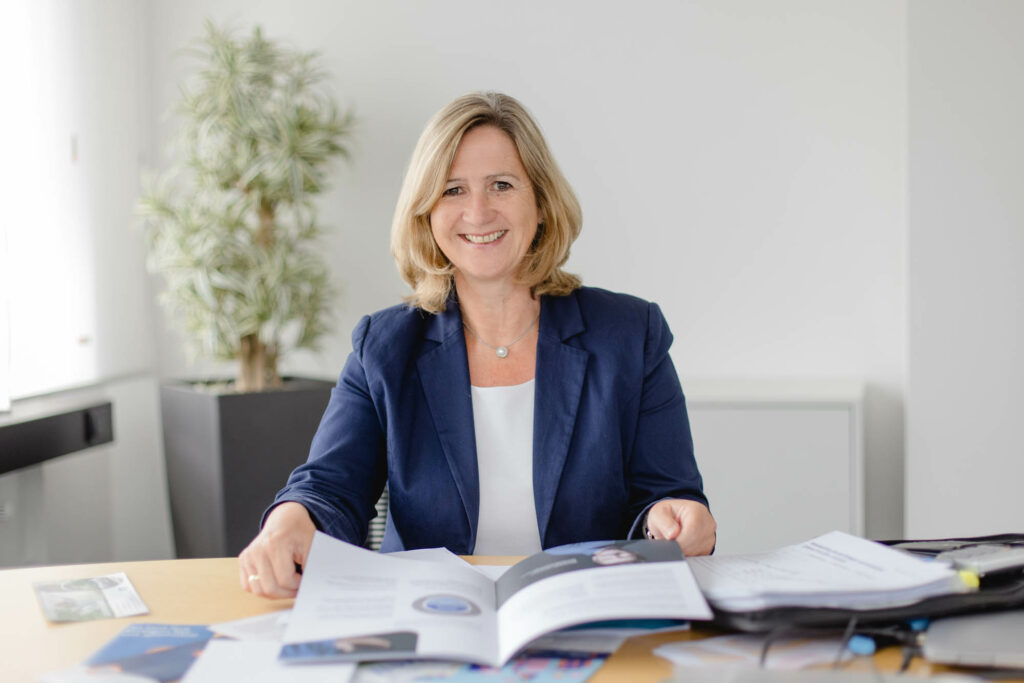 Eine blonde Frau in einem blauen Blazer sitzt an einem Tisch. Vor ihr liegt eine öffene Aktenmappe. Sie lacht.