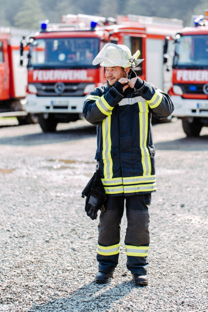 Ein Feuerwehrmann in Dienstkleidung. Er macht sich den Helm zu. Er lacht. Dahinter stehen die Feuerwehrautos.