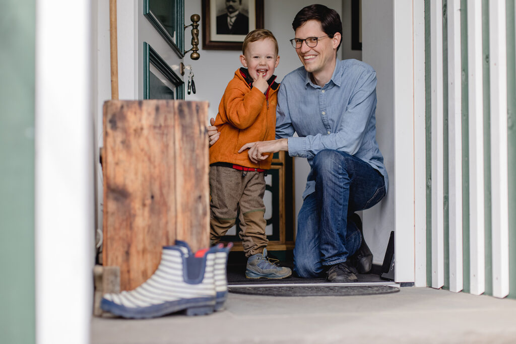 Mann und ein Kind in orangener Jakce stehen in einem Türrahmen. Links stheen Gummiestiefel im Bild.