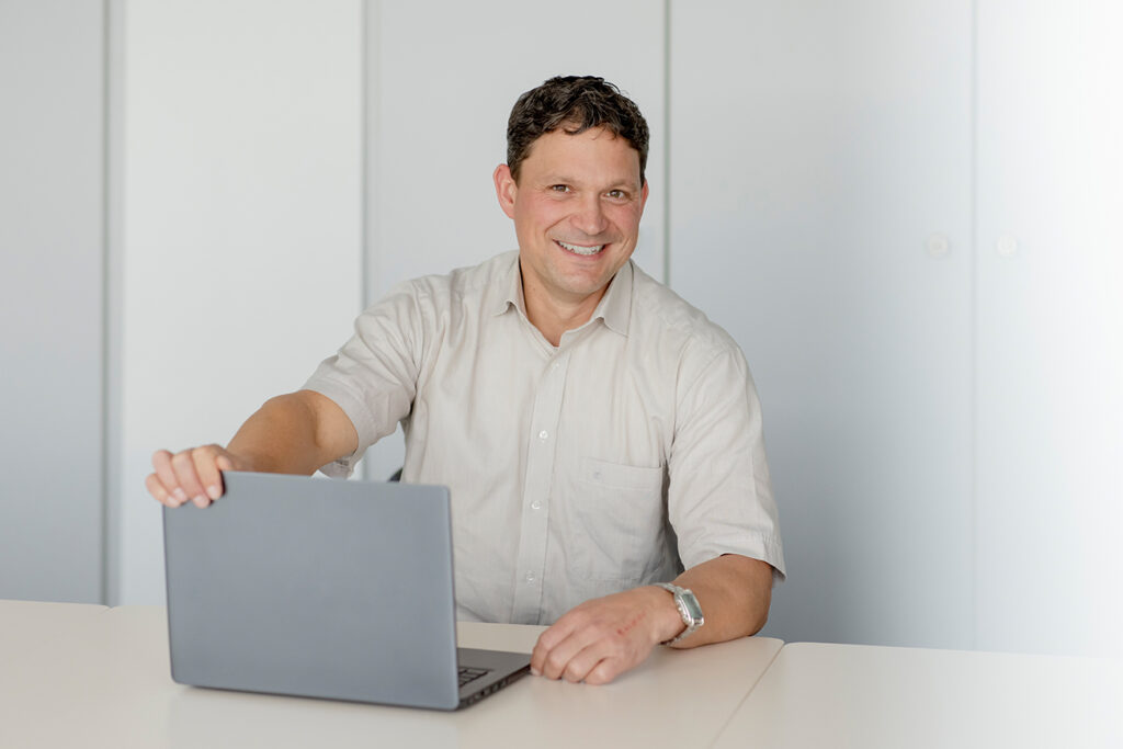 Mann mit kurzen brauen Haaaren sitzt an einem Tisch. Er trägt ein beiges Hemd und hat einen laptop vor sich.
