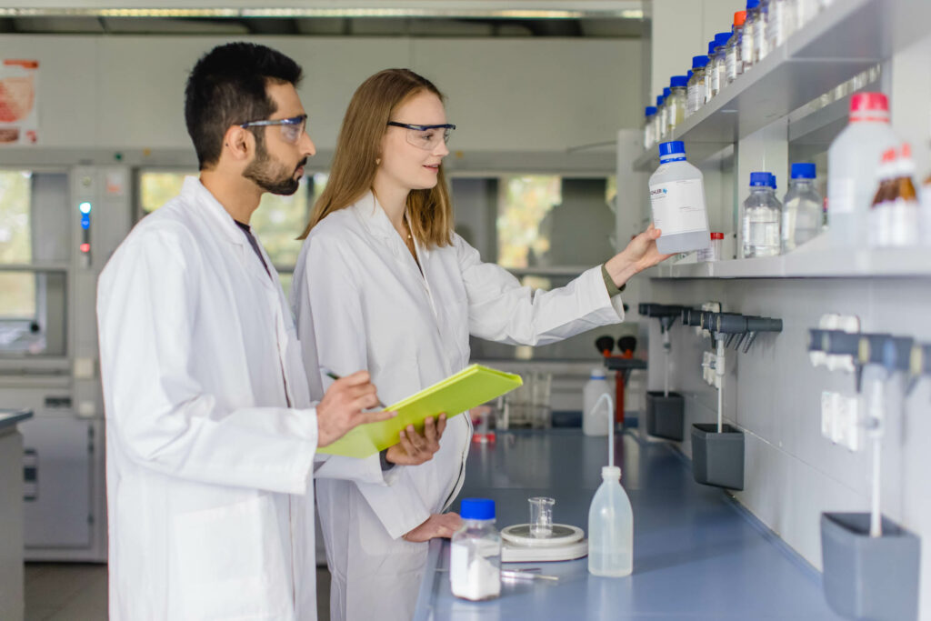 Ein Mann und eine Frau sehen in weißen Kitteln in einem Labor vor einem Regal. Sie tragen Schutzbrillen.