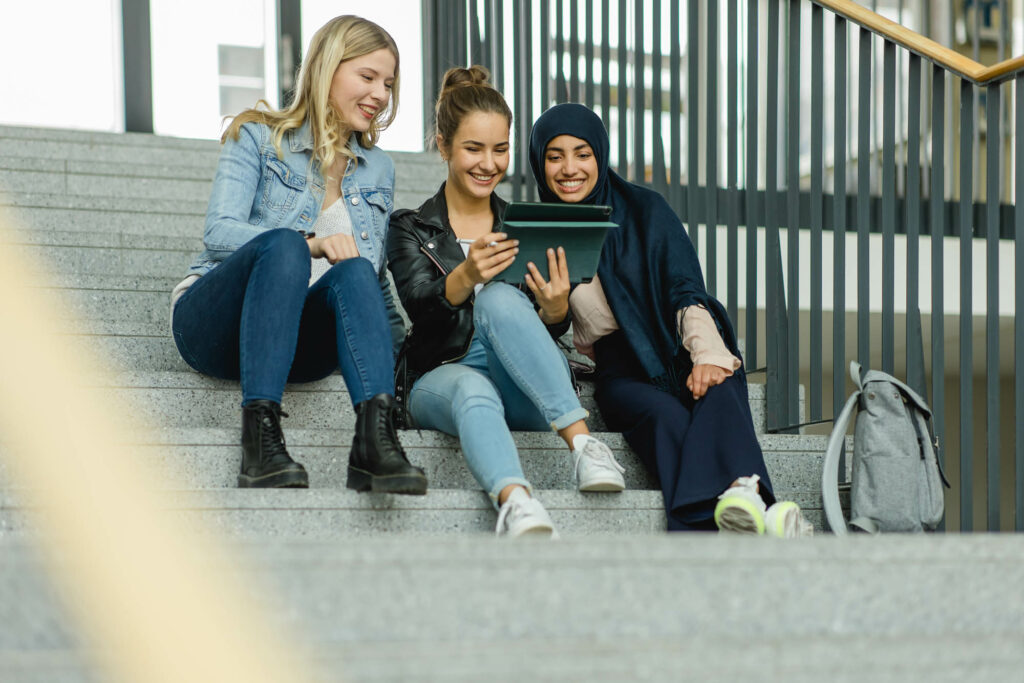 Drei Studentinnen sitzen auf Treppenstufen und schauen auf ein Tablet.