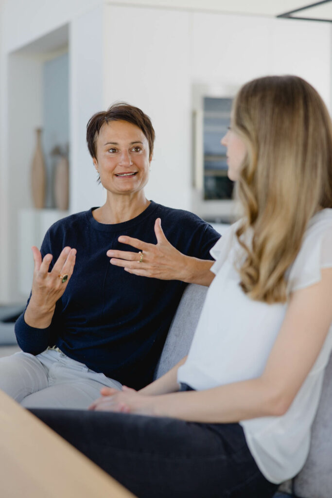 Eine Frau mit bloden langen Haaren und eine Frau mit kurzen dunklen Haaren sitzen auf einer hellen Couch und untehrlaten sich.