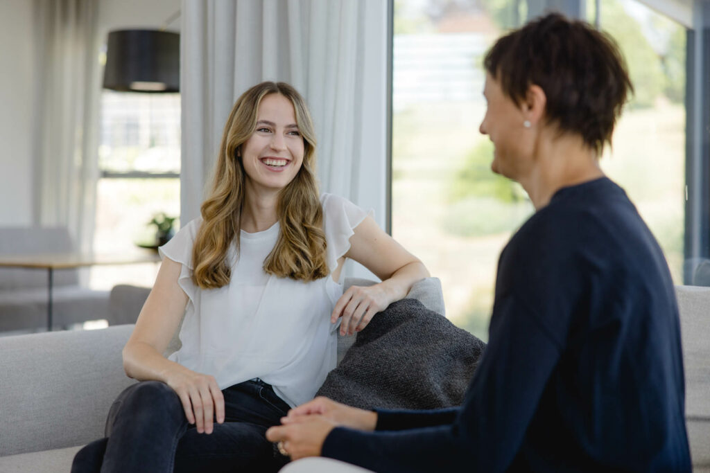Eine Frau mit bloden langen Haaren und eine Frau mit kurzen dunklen Haaren sitzen auf einer hellen Couch. Sie lachen.