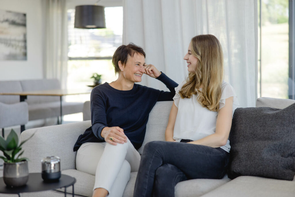 Eine Frau mit bloden langen Haaren und eine Frau mit kurzen dunklen Haaren sitzen auf einer hellen Couch. Sie lachen.