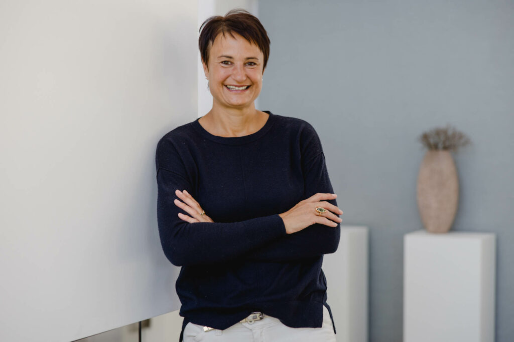 Portrait von einer Frau mit kurzen dunklen Haaren in einer dunklen Bluse und mit verschränkten Armen. Sie lacht.