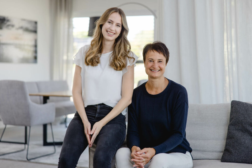 Eine Frau mit bloden langen Haaren und eine Frau mit kurzen dunklen Haaren sitzen auf einer hellen Couch. Sie lachen.