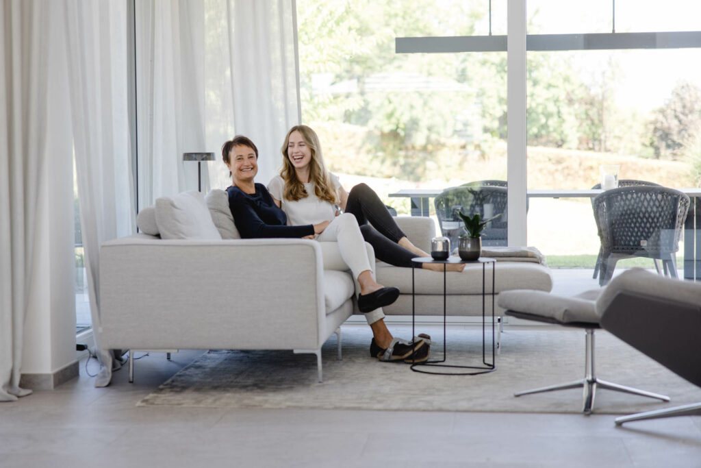 Eine Frau mit bloden langen Haaren und eine Frau mit kurzen dunklen Haaren sitzen auf einer hellen Couch. Sie lachen.