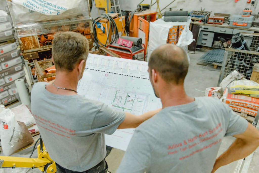 Zwei Männer in grauen T-Shirts schauen von oben auf eine Bauplan, den sie in den Händen halten.
