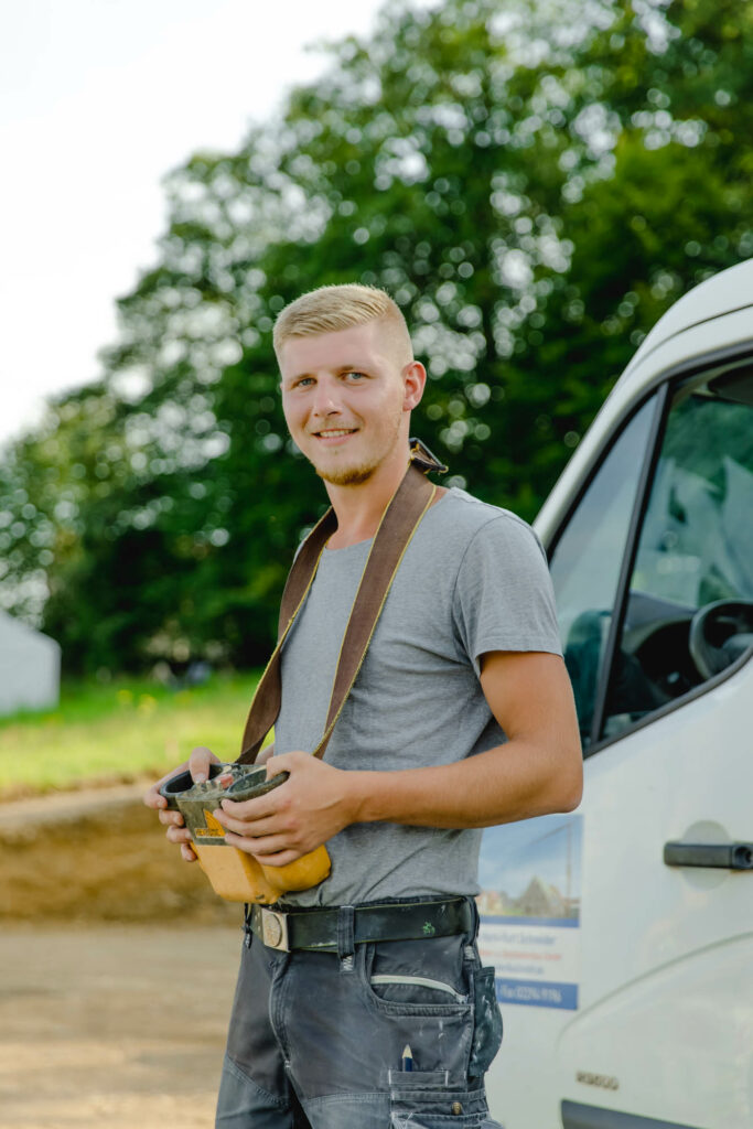 Ein blonder Mann hält eine Kranfernbedienung in der Hand. Rechts hinter ihm steht ein weißer Transporter. Er lacht.