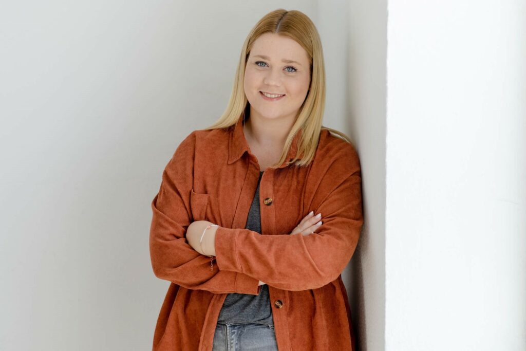 Frau mit blonden Haaren und oranger Jacke lehnt an einer weißen Wand.