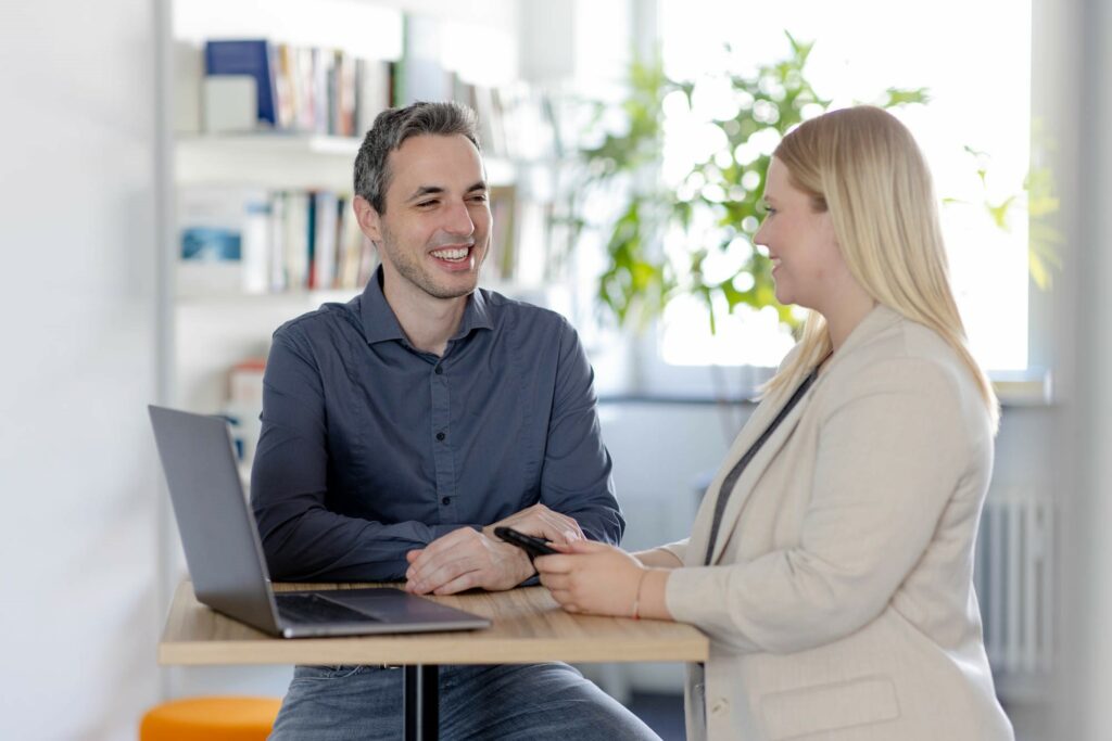 Mann und Frau stehen an einem Tisch. Vor ihnen ein Laptop und unterhalten sich.