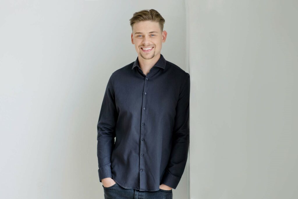 Mann mit kurzen Haaren, dunklem Hemd lacht und lehnt an einer weißen Wand.