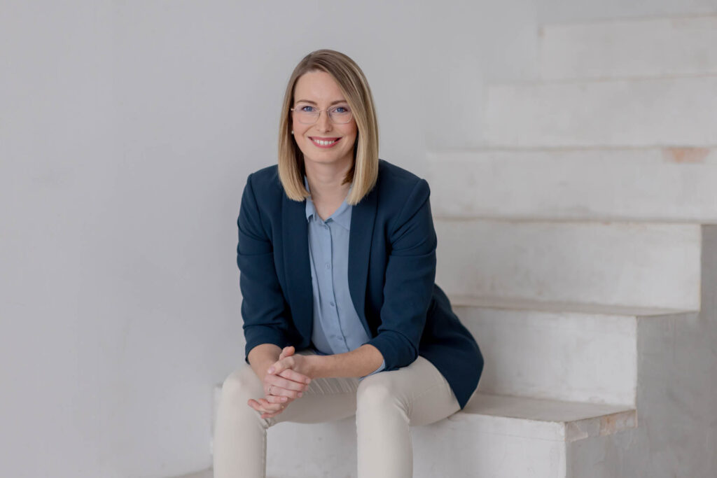 Frau mit blonden, schulterlangen Haaren sitzt auf einer Treppe und lacht, Sie trägt einen braunen Blazer und eine helle Hose.