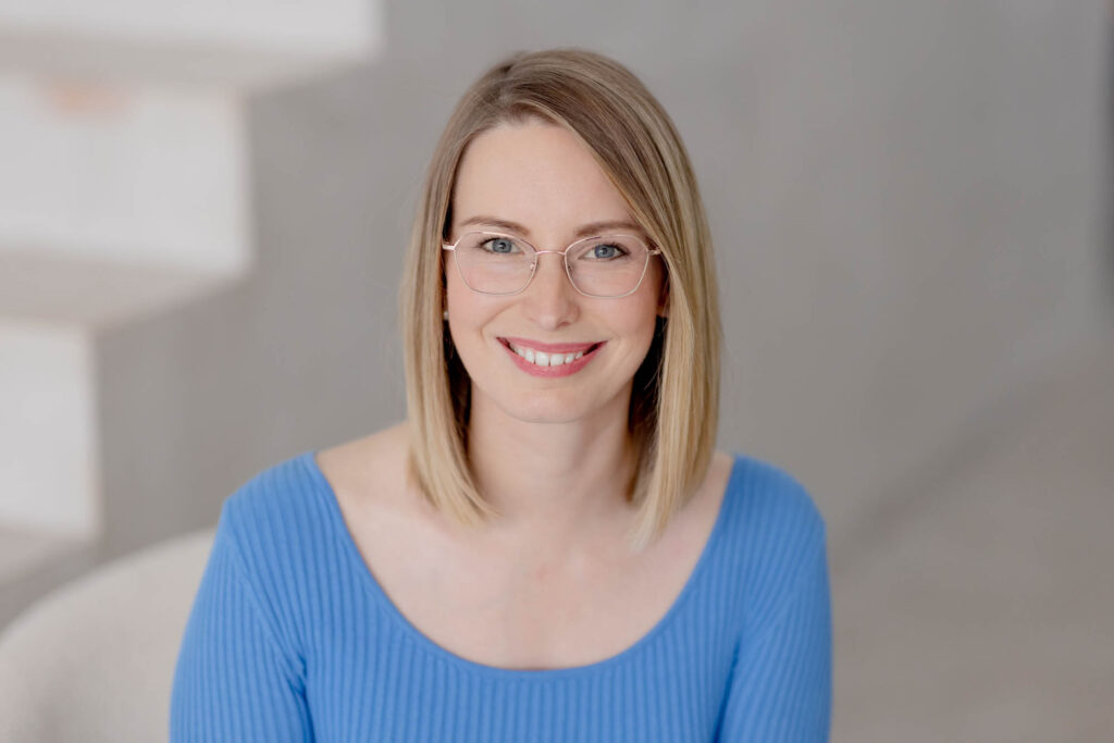 Portrait von einer Frau mit blonden Haaren in blauem Pulli und Brille.