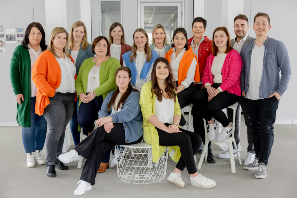 Gruppenfoto eines Unternehmens mit vielen bunten Farbakzenten. Zwei Personen sitzen vorne. Alle lächeln.